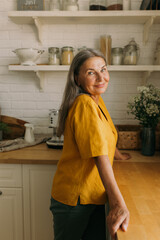 Smiling mature female posing in modern home kitchen. Happy middle-aged woman standing at tabletop...