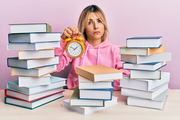 Young caucasian woman holding alarm clock sitting on the table with books skeptic and nervous, frowning upset because of problem. negative person.