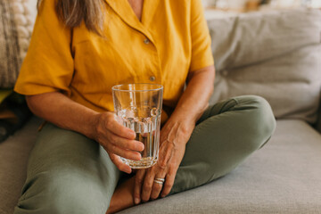 Glass of water in female hand. Mature woman sitting on sofa and holding glassful with beverage....