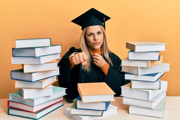 Young caucasian woman wearing graduation ceremony robe sitting on the table punching fist to fight, aggressive and angry attack, threat and violence