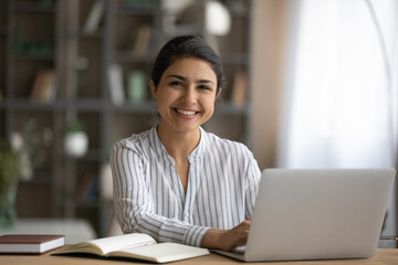 Portrait of happy millennial Indian female student sit at desk study distant on computer. Smiling...