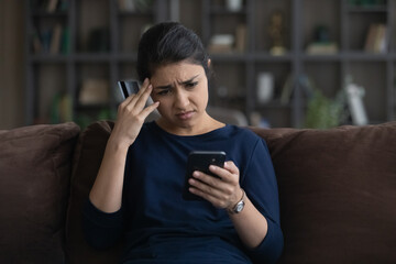 Unhappy young Indian woman distressed with payment problems shopping online from home on smartphone...