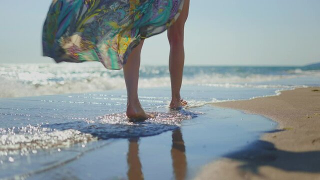 Tracking footage of female legs stepping into waves washing sandy beach. Glittering water surface. Summer vacation at sea, relaxing on beach.