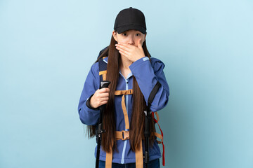 Obraz na płótnie Canvas Young Chinese girl with backpack and trekking poles over isolated blue background covering mouth with hand