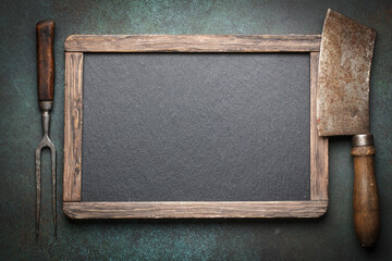 Vintage menu blackboard with old meat cleaver and fork on dark background with copy space