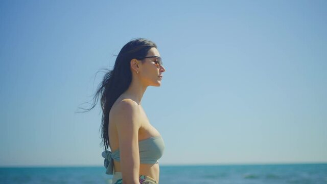 Attractive Caucasian brunette walking around ocean coast in sunny day. Waist up side tracking shot. Summer vacation at sea, relaxing on beach.