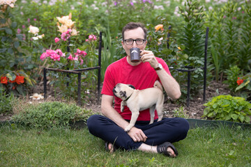 Naklejka na ściany i meble A man with glasses sits on a lawn in a blooming garden and drinks tea from a mug with a painted mustache. A pug puppy is on the man's lap.