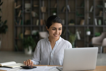 Happy young Indian woman sit at desk pay bills online on laptop, calculate expenses expenditures....