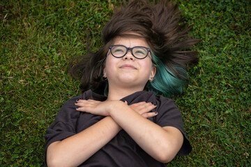 A teenage girl in glasses with multi-colored hair and with earrings lies on the lawn with her arms crossed over her chest. She looks up to the sky and smiles. 