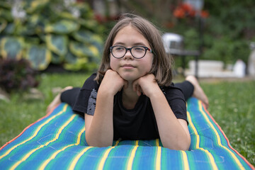 A teenage girl with glasses lies on a mattress on a lawn in a summer garden and thinks about something