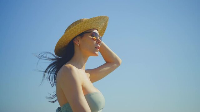 Walking attractive mid adult brunette against blue clear sky. Wearing sunglasses and straw hat. Summer vacation at sea, relaxing on beach.