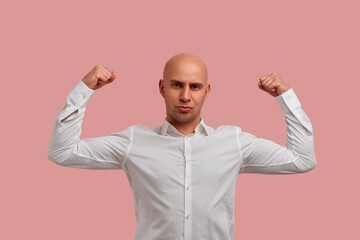 Fototapeta premium Strong bald man with bristle posing raises hands and shows muscles, his strength and power, feels proud about his achievements in gym. Dressed in white shirt, isolated over pink background.