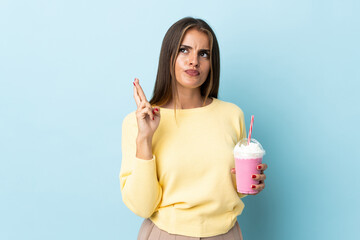 Young Uruguayan woman with strawberry milkshake isolated on blue background with fingers crossing and wishing the best