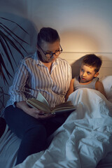 grandmother in glasses and a white shirt reads a book to her grandson lying on the bed in a white children's room