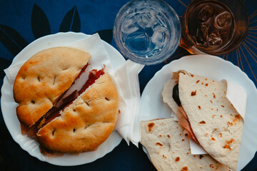 Traditional Italian food. Crispy panini and piadina sandwiches with prosciutto (speck), cheese, lettuce and vegetables with fresh drinks on a table with a blue tablecloth. Healthy European snack.