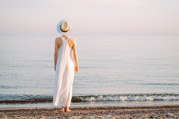 Young slim woman wearing white long dress and hat walk alone at the beach or ocean or sea....