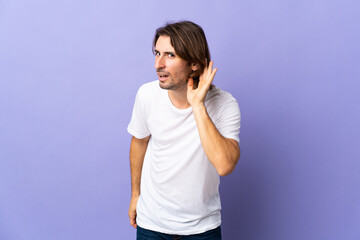 Young handsome man isolated on purple background listening to something by putting hand on the ear