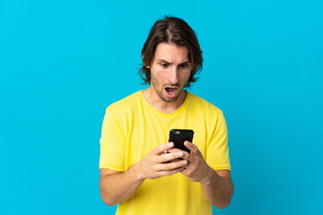 Young handsome man isolated on blue background looking at the camera while using the mobile with surprised expression