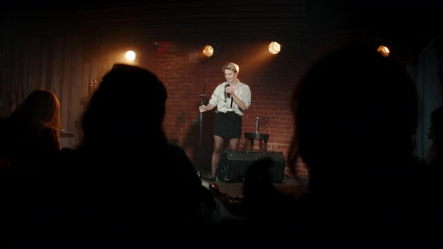 Young Caucasian female comedian performing her stand-up monologue on a stage of a small venue. Shot with ARRI Alexa Mini LF with 2x anamorphic lens
