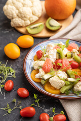 Vegetarian salad of cauliflower cabbage, kiwi, tomatoes, microgreen sprouts on black concrete background. Side view, selective focus.