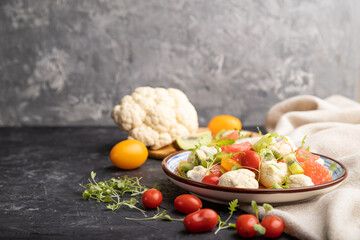 Vegetarian salad of cauliflower cabbage, kiwi, tomatoes, microgreen sprouts on black concrete background. Side view, copy space.
