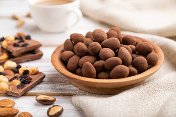 Almond in chocolate dragees in wooden plate and a cup of coffee on white wooden background. Side view, selective focus.