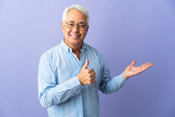 Middle age Brazilian man isolated on purple background holding copyspace imaginary on the palm to insert an ad and with thumbs up