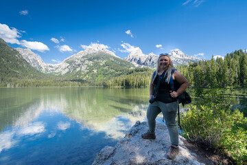 Proud blonde woman hiker stands on a rock on the shoreline of Taggart Lake, in Grand Teton National...