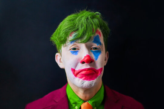 Man in mime makeup cosplay with green hair and a red suit an orange tie and a green shirt. Clown sad