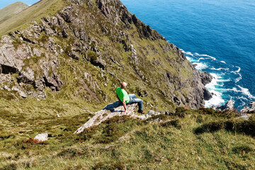 Bald male tourist in green shirt sitting on the edge of a rock. Achill island, county Mayo,...