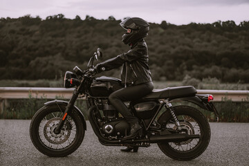Fototapeta na wymiar Portrait of confident motorcyclist woman in motorcycle helmet. Young driver biker looking away outdoors alone on highway. Ready for trip. Cafe racers, motorbike aesthetics and vintage design concept.