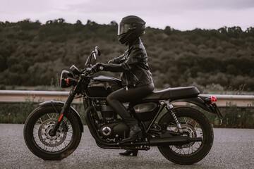 Portrait of confident motorcyclist woman in motorcycle helmet. Young driver biker looking away outdoors alone on highway. Ready for trip. Cafe racers, motorbike aesthetics and vintage design concept.