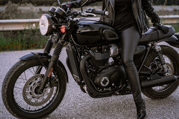 Fototapeta na wymiar Legs of stylish motorcyclist woman sitting on classic bike. Black retro-styled motorcycle. Details of vintage design of brand-new motorbike.