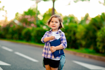 a schoolgirl is standing on the road in a plaid shirt and a medical mask, with a school backpack and a diary in her hands