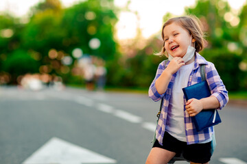 Fototapeta premium a schoolgirl is standing on the road in a plaid shirt and a medical mask, with a school backpack and a diary in her hands