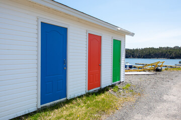 Fototapeta na wymiar A row of colorfully painted solid doors of blue, red, and green. The exterior wall is white vinyl siding. The sky is blue in the background and the storage units are sitting on gravel with blue ocean.