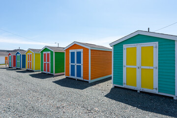 Fototapeta na wymiar A row of small colorful painted huts or sheds made of wood. The exterior walls are colorful with double wooden doors. The sky is blue in the background and the storage units are sitting on gravel.