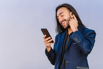 Young Hispanic Latin business man with long hair using wireless earphons with his smart phone, isolated. gray wall background. copy space