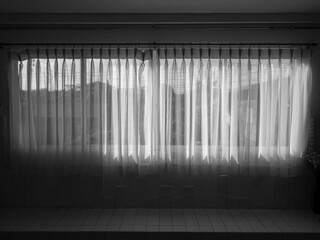 White transparent curtain at the window in the dark room. Large horizontal window with closed white fabric curtain.