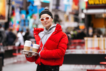 Urban portrait of stylish young girl on New York street with coffee to go. Casual modern woman wearing trendy sunglasses, red jacket and street style clothes over Time Square, NYC downtown background
