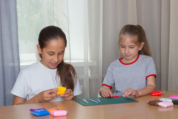 Naklejka premium Two girls sisters of school and preschool age sculpt from colored clay at home. There is a lot of colored clay on the table.