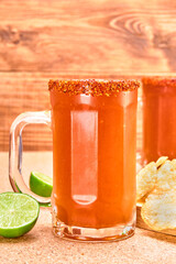 mexican style beer mug (Chelada/Michelada) with lime, hot sauce and chamoy, frosted with chili powder. accompanied by potato chips on a cork surface and wooden background. close up view