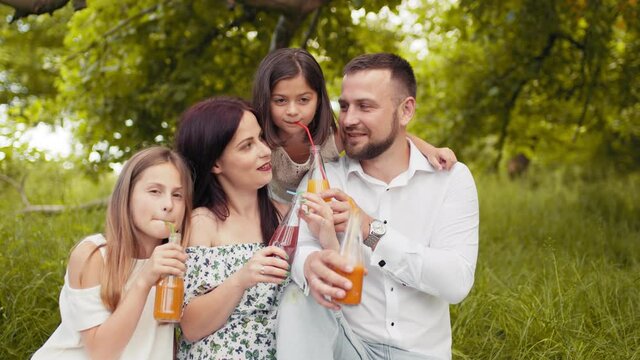 Smiling father, mother and two cute daughters drinking fresh juice at green garden while having summer picnic. Concept of family, leisure and relaxation. Family spending cheerfully free time on nature