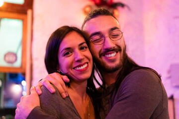 young hispanic latin man and woman hugging, friends in a bar laughing looking at the camera