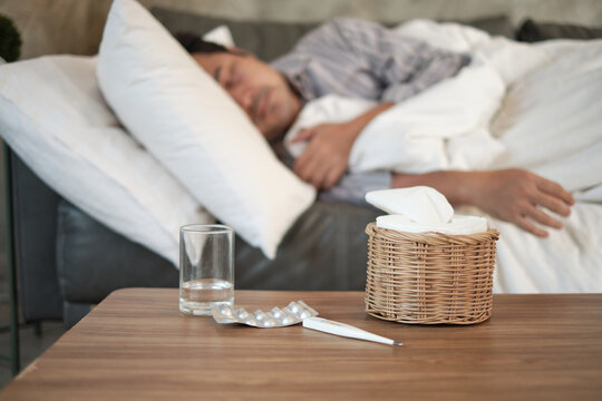 Asian man is absent from work and sick leave because of illness, takes temperature by thermometer, takes medicine, and sleeps in blankets on sofa at home, tissues, and capsule pills on the table.