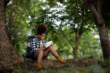  American black female teenager hiking backpacker sitting nex to big tree in the natural forest and take note of a notebook