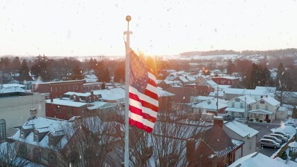 American flag at winter sunrise, sunset as snowflakes fall in pretty town scene. Aerial view. - Powered by Adobe