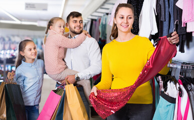Young happy cheerful mother choosing new dress in shop while father with daughters standing behind
