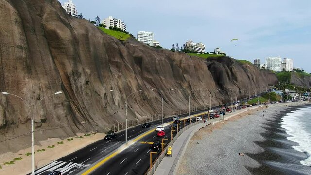 Boom job down 4k aerial video of the Miraflores coast in Lima, Peru, in a summer sunny day. People are paragliding and relaxing on the cobblestone beach looking at the powerful pacific ocean waves.