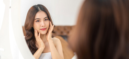 Obraz premium Smiling of young beautiful pretty asian woman clean fresh healthy white skin looking at mirror.asian girl touching on her face with hand and applying cream at home.spa and beauty concept